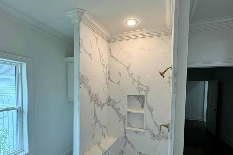 Shower in a marble look slab framed by white crown molding and trim, beside a window and pale gray walls