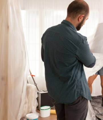 Young man covering lamp with plastic sheet before start renovating living room. Home during renovation, decoration and painting. Interior apartment improvement maintenance. Roller, ladder for house repair